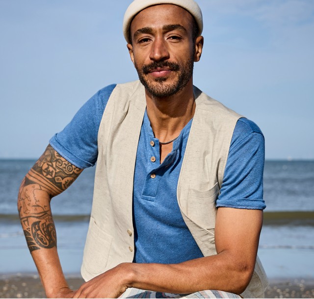Das Bild zeigt einen Mann bekleidet mit einem blauen T-Shirt und einer hellen Weste darüber; hinter ihm sieht man die Wellen des Ozeans.