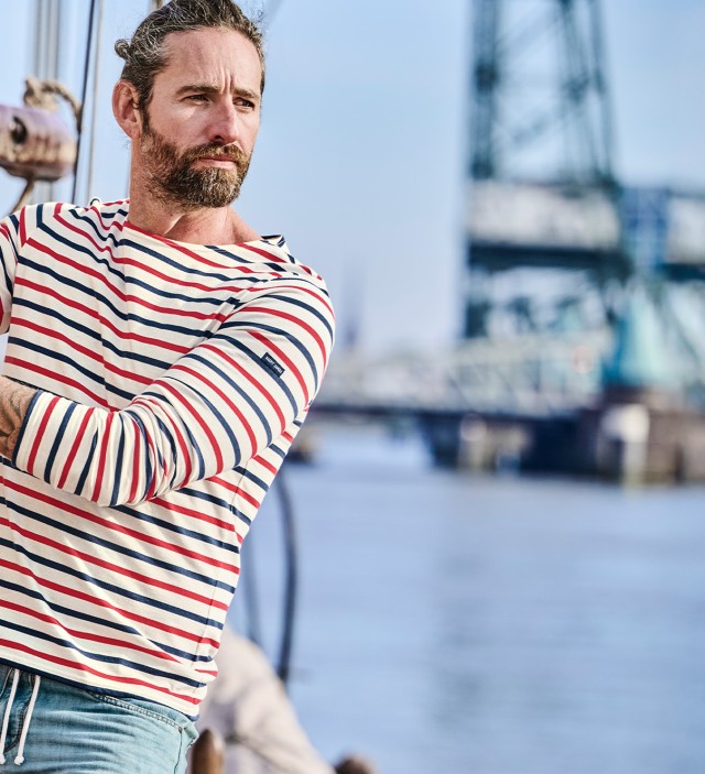 Mann bekleidet mit einem bunt  gesteiftem Langarmshirt und einer hellen Jeans steht auf einem Segelboot und hält sich am an einem Seil fest.