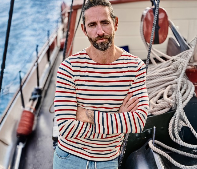 Mann bekleidet mit einem bunt gestreiftem Langarmshirt  steht mit verschränkten Armen auf einem Segelboot und blickt direkt in die Kamera