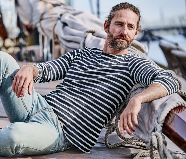 Mann bekleidet mit einem blau gestreiftem Langarmshirt  liegt entspannt auf dem Deck eines  Segelbootes und blickt in die Ferne.
