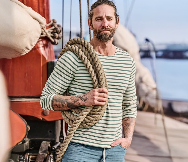Mann bekleidet mit einem grün gestreiftem Langarmshirt  und einer Jeans steht lässig auf dem Deck eines  Segelbootes. Über seiner Schulter hängt ein dickes Tauseil.