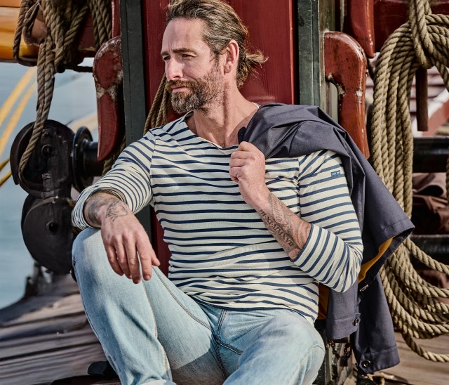 Mann bekleidet mit einem blau-weiß gestreiftem Langarmshirt  sitzt entspannt auf dem Deck eines  Segelbootes. Über seiner Schulter trägt er ein blaues Blouson