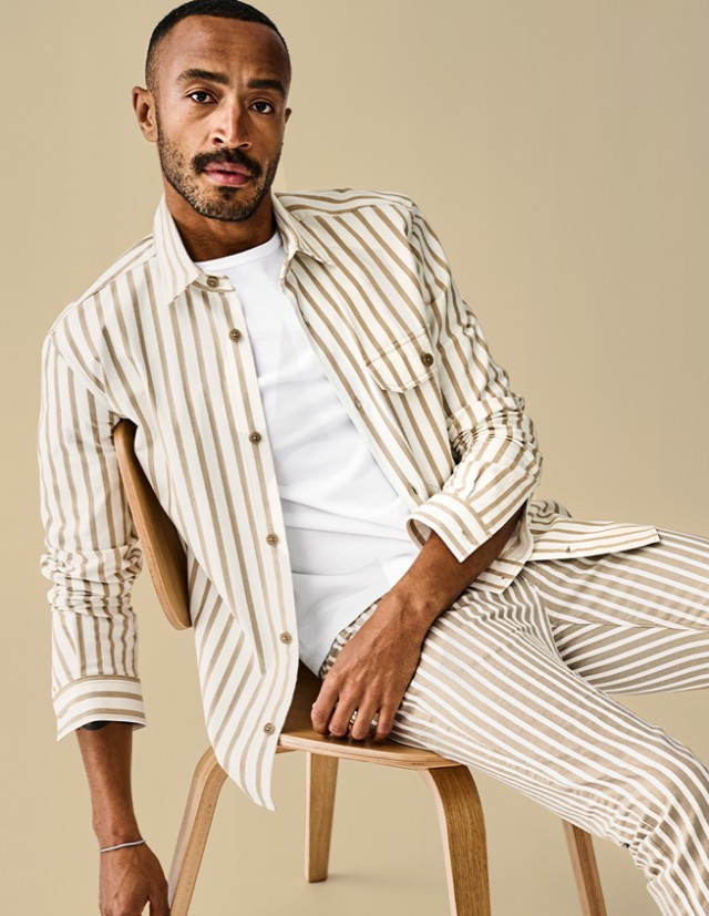 Mann sitzt vor einem beigem Hintergrund lässig auf einem Stuhl und blickt in die Kamera. Er trägt ein beige-weiß gestreiftes Hemd, sowie eine passend gestreifte Hose dazu.