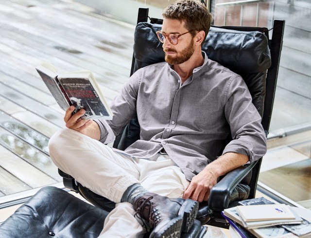 Mann trägt ein graues Hemd, sitzt entspannt in einem Ledersessel und liest ein Buch