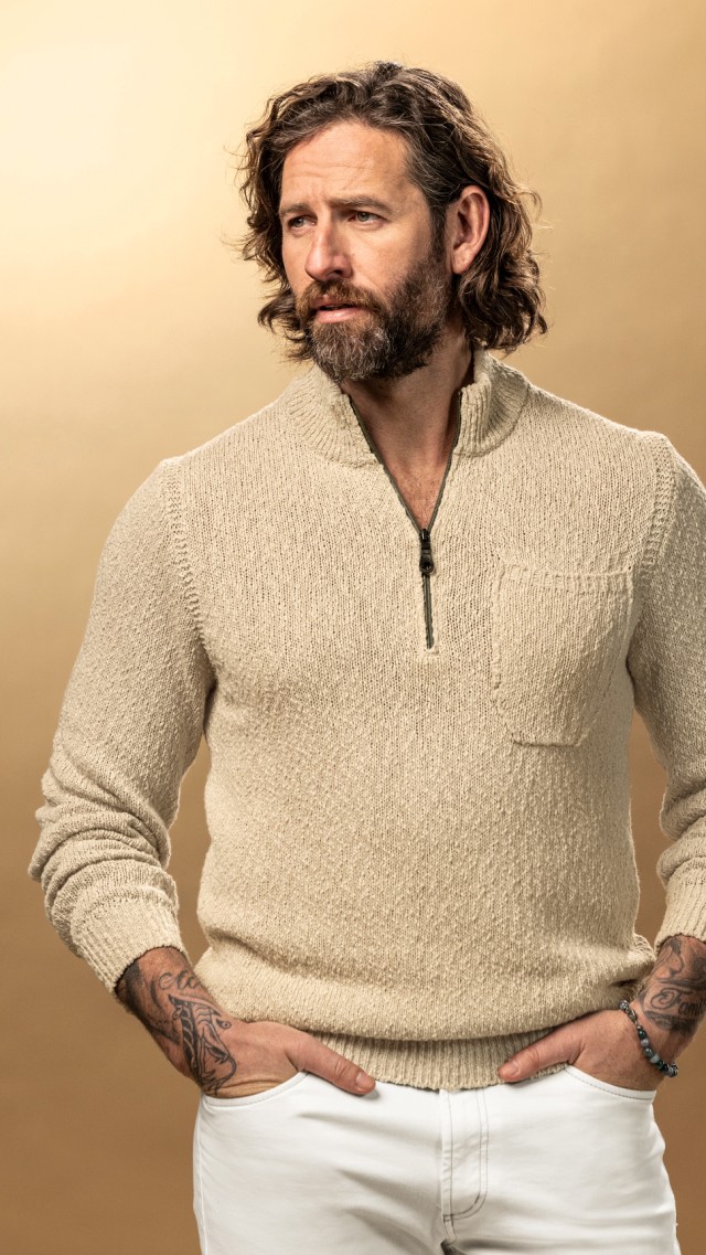Mann bekleidet mit einem beigefarbenen Strickpullover und einer weißen Jeans