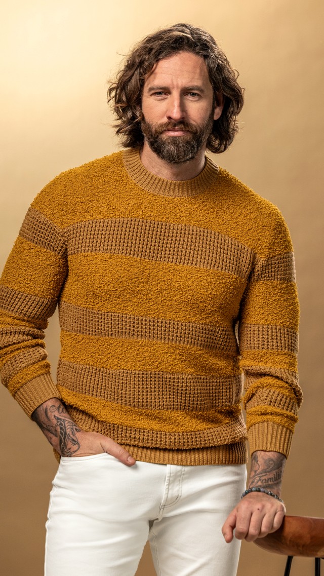 Mann mit einem orangefarbenen Strickpullover und weißer Hose blickt in die Kamera