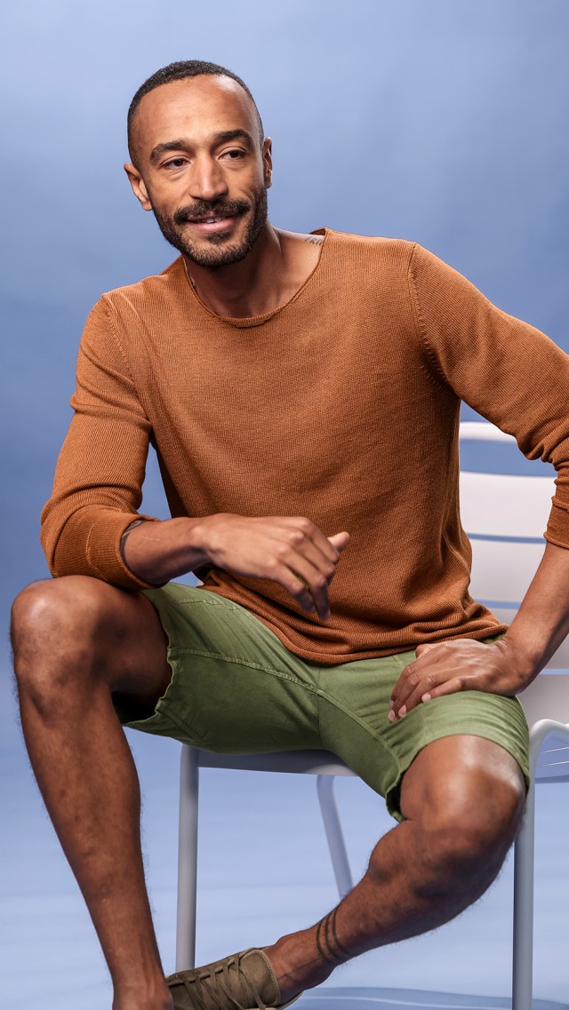 Ein Mann trägt einen rot-braunen Strickpullover und eine oliv farbene Hose und sitzt auf einem Stuhl.
