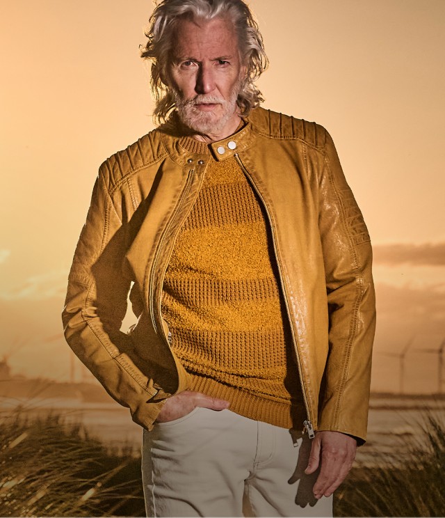 Mann mit einer gelben Lederjacke und einer hellen Hose steht am Meer vor einem spektakulären Sonnenuntergang