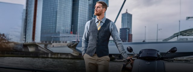 Ein Mann bekleidet mit einem hellblauen Hemd trägt eine dunkelblaue Weste darüber und hält eine Hand an einer Vespa