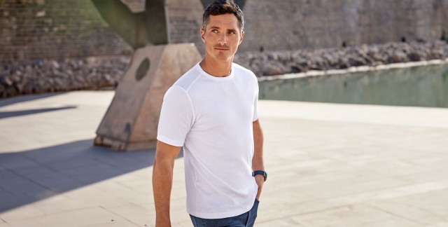 Mann im weißen T-Shirt und Jeans spaziert in sonniger, urbaner Umgebung.