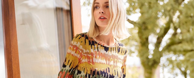 Eine Frau trägt einen farbenfrohen Pullover mit abstraktem Muster. Sie steht neben einer Glastür mit Blick auf die Natur im Hintergrund.