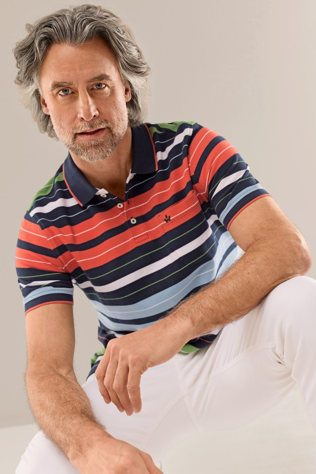 Ein grauhaariger Mann trägt ein gestreiftes Poloshirt in Blau, Grün, Rot und Hellblau und steht entspannt vor hellem Hintergrund.