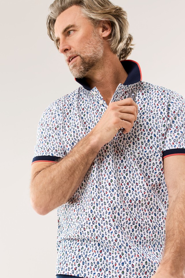 Ein grauhaariger Mann trägt ein weißes Poloshirt mit buntem, kleinteiligem Muster und aufgestelltem Kragen, während er lässig zur Seite blickt.