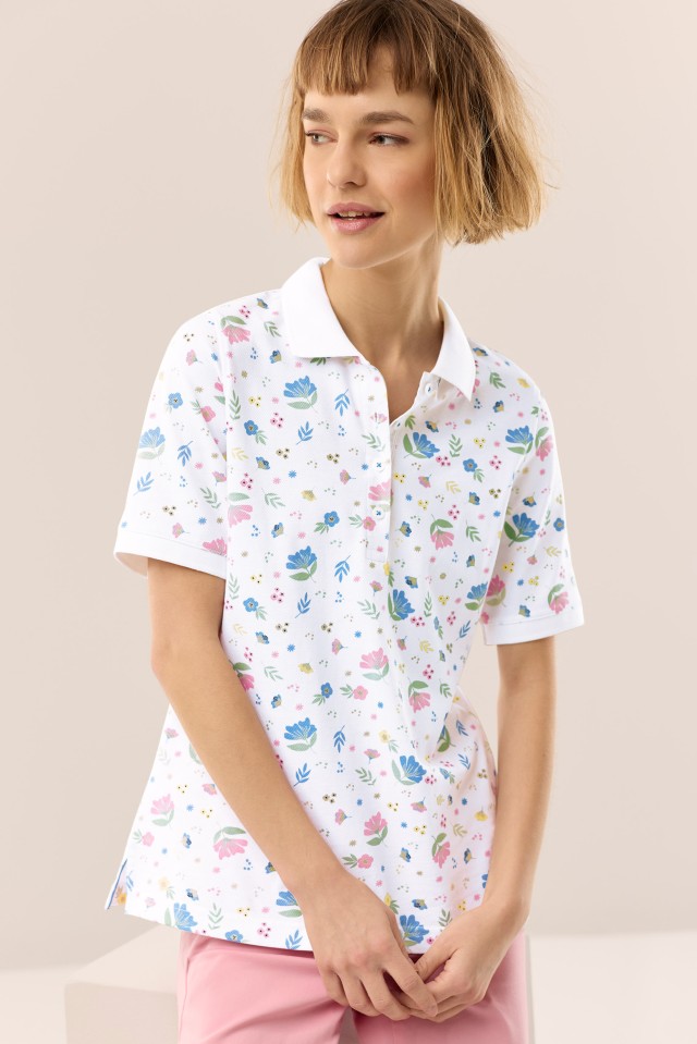 Eine blonde Frau trägt ein weisses Poloshirt mit floralen, farbigen Akzenten und steht vor neutralem Hintergrund.