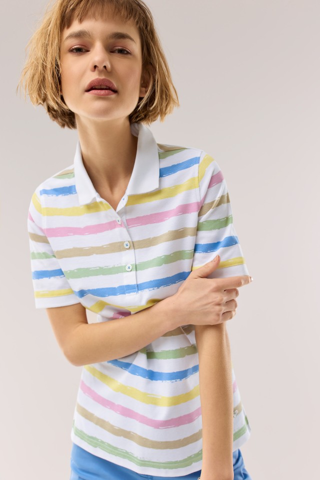 Eine blonde Frau trägt ein weisses Poloshirt mit farbigen Streifen und schaut in die Kamera.