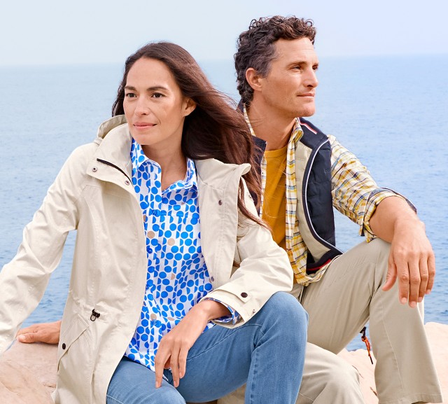 Eine Frau und ein Mann sitzen auf einem Felsen am Meer und tragen leichte Outdoorkleidung.