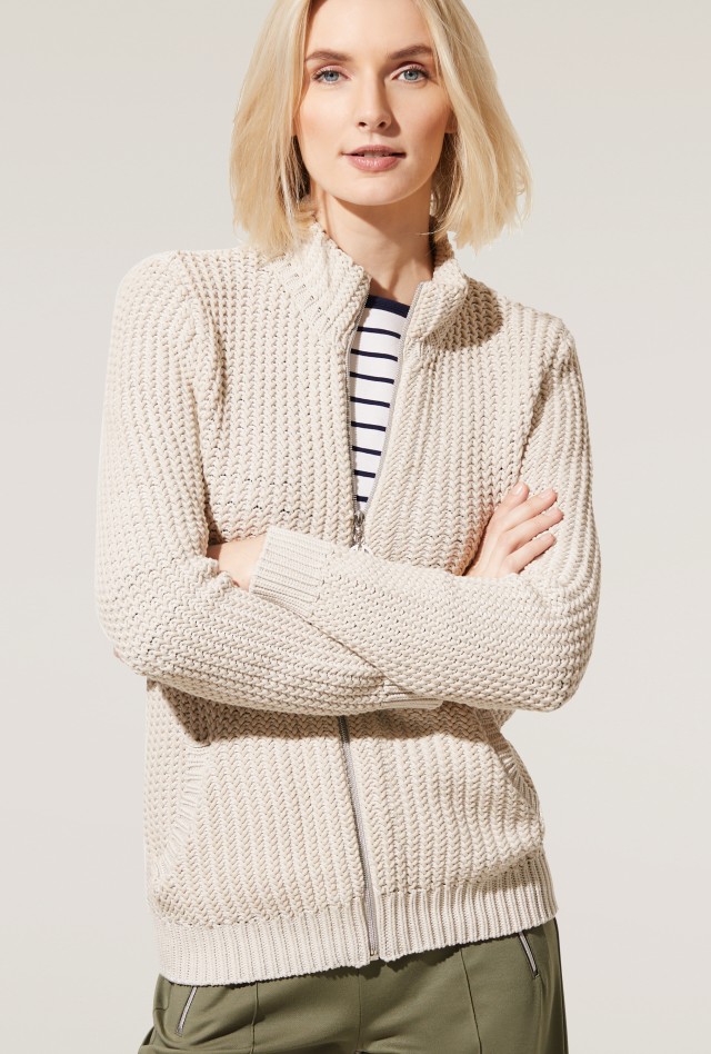 Frau in einer beigen Strickjacke und gestreiftem Shirt steht vor einem neutralen Hintergrund.