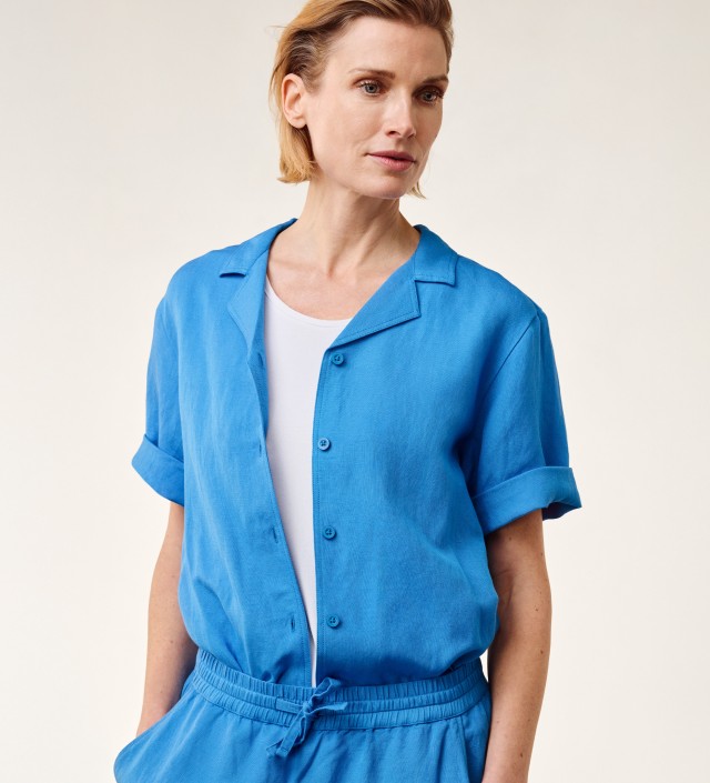 Blonde Frau in einer blauen Leinenbluse und -hose vor einem hellen Hintergrund schaut zur Seite