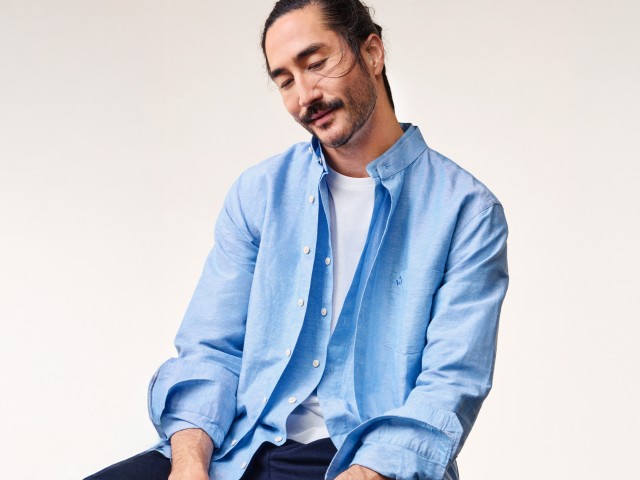 Mann in hellblauem Leinenhemd und weißem Shirt vor einem hellen Hintergrund schaut nach unten