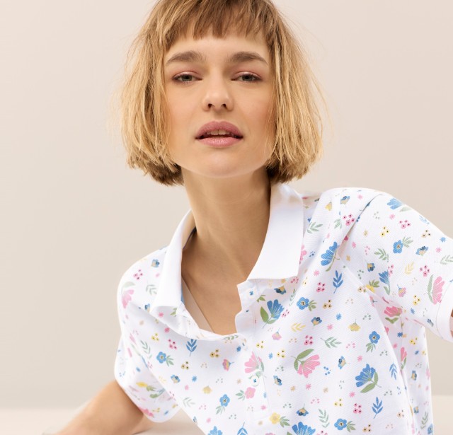 Eine blonde Frau trägt ein weisses Poloshirt mit floralen, farbigen Akzenten und schaut in die Kamera.