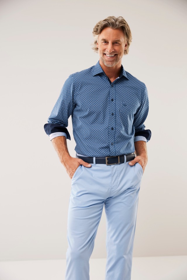 Mann in blauem, gemusterten Hemd und hellblauer Hose lächelt in die Kamera.
