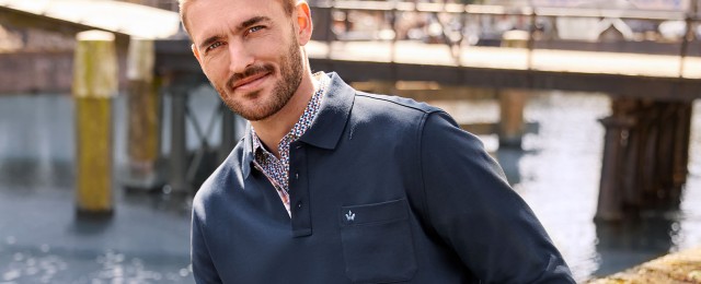 Ein Mann steht an einem Gewässer vor einer Brücke und trägt ein dunkelblaues Polohemd mit einer Brusttasche. Darunter trägt er ein Hemd mit buntem Muster.