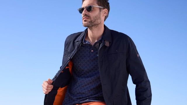 Mann in schwungvoller Pose trägt eine Sonnenbrille, einen marineblauem Blouson und Polo-Shirt
