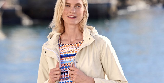 Frau in einem gemusterten Oberteil und einer hellen Jacke steht am Meer und schaut in die Kamera.