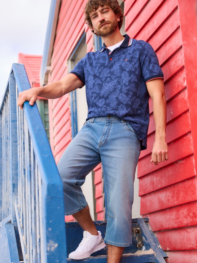 Ein Mann mit lockigen Haaren steht auf einer blauen Treppe vor einem roten Gebäude, trägt ein blau gemustertes Polohemd, lange Jeansshorts und weiße Turnschuhe.