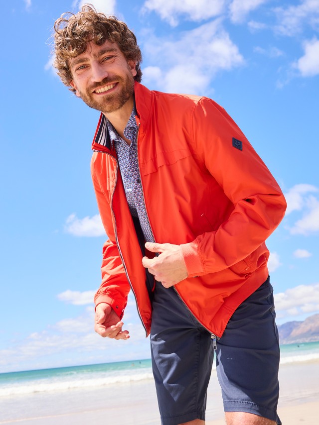 Ein lachender Mann steht am Strand und trägt eine leuchtend orangefarbene Jacke über einem gemusterten Hemd sowie dunkelblaue Shorts.