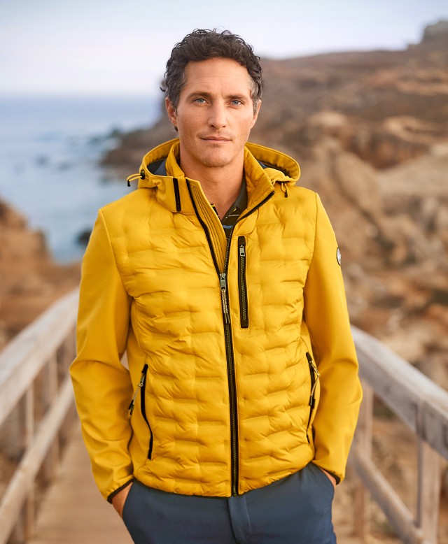 Mann steht auf einer Brücke am Meer und trägt eine gelbe Jacke.