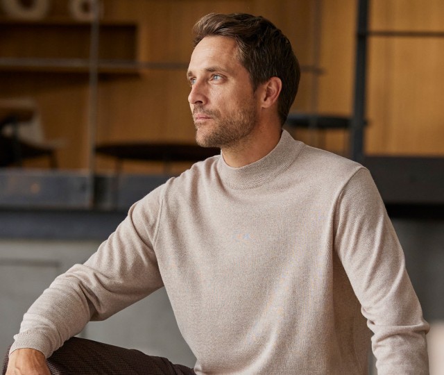 Herr sitzt und trägt einen beigen Merino Rolkragen Pullover.