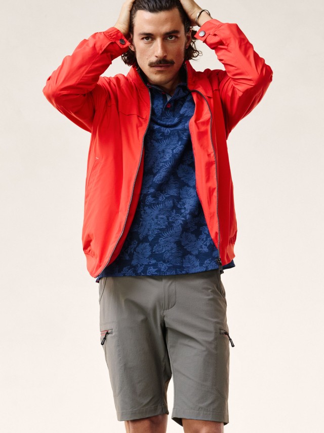 Ein Mann trägt einen roten Blouson, ein gemustertes Polo-Shirt, Bermudas in khaki und streicht sich mit beiden Händen durch die Haare.
