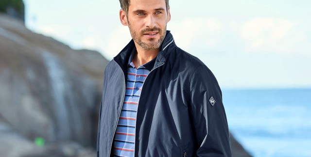 Mann in marineblauem Blouson und gestreiftem Polo-Shirt steht an einer Felsküste