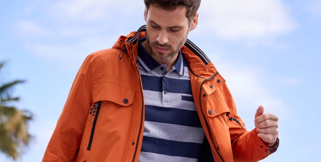 Mann in einem blau-gestreiften Polo und orangener Jacke steht am Meer.