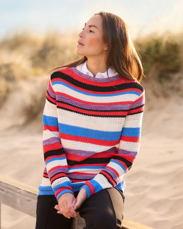 Frau sitzt auf einem Holzzaun am Strand und trägt einen gestreiften Pullover.