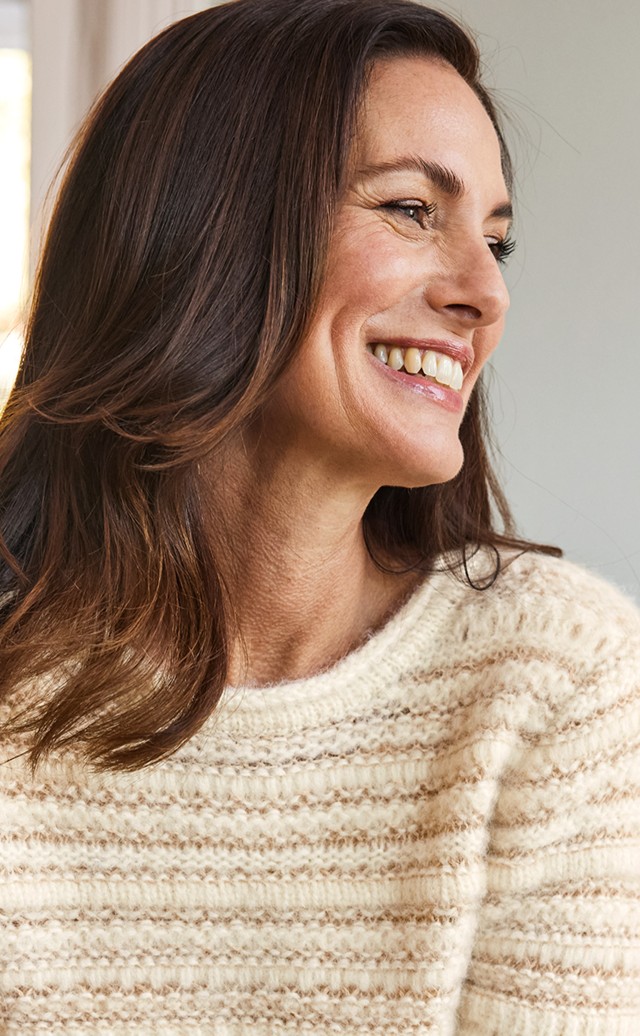 Frau im kuscheligen beigefarbenen Wollpullover steht lächelnd im Wohnbereich.