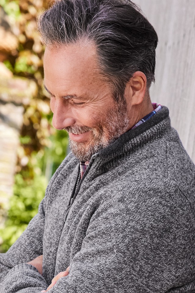Mann im graumelierten Pullover lehnt lässig an einer Holzwand im Garten.