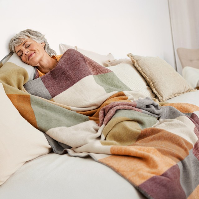Frau kuschelt sich auf einer Couch in eine Wohndecke in sommerlichen Farben.