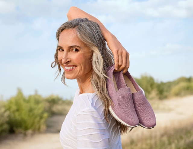 Die grauhaarige Frau genießt das Leben und steht bei sonnigem Wetter mit ihren Hallux-Slipper im frühlingshaften Mauveton in der Hand in einer sommerlichen Landschaft.