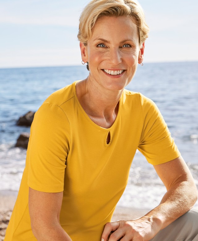 Frau trägt ein gelbes Shirt und sitzt am Meer.
